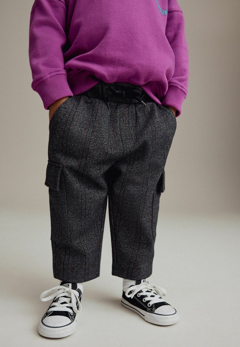 Child wearing gray pinstriped pants, black and white sneakers, white socks, and a purple sweatshirt with hands in pockets.