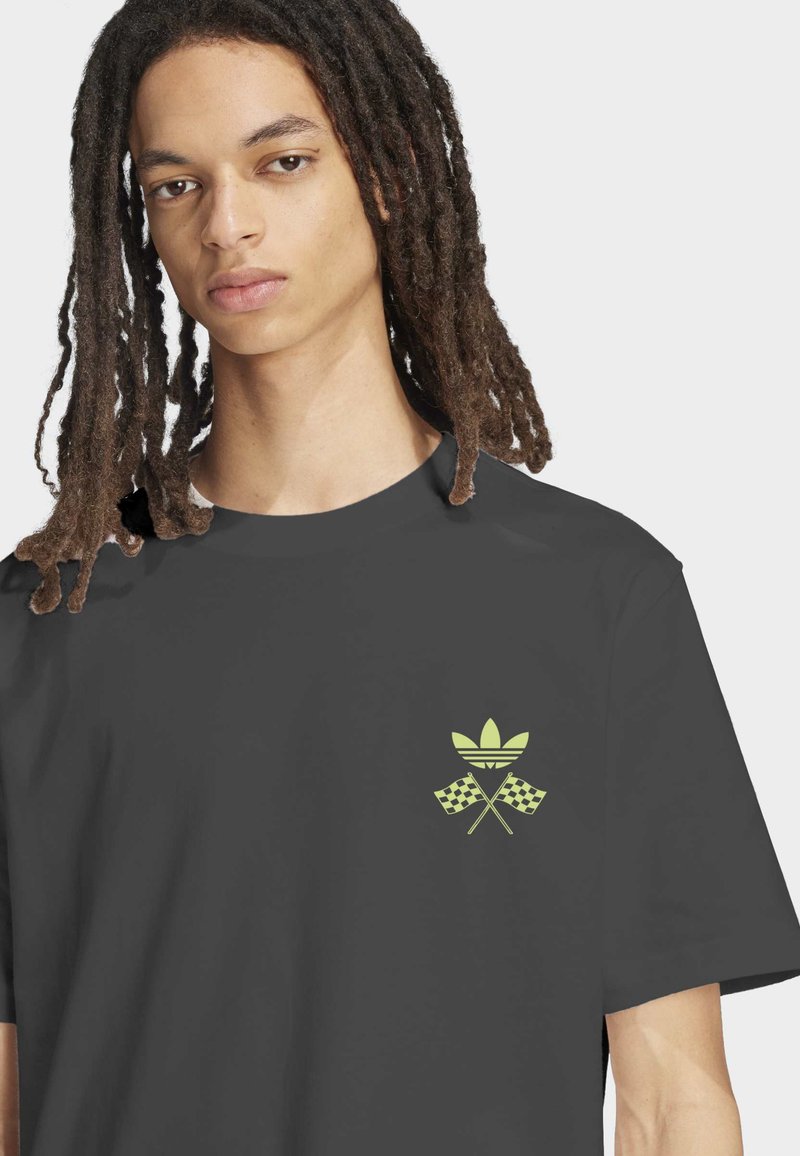 Jongere met lange dreadlocks, gekleed in een donkergroen T-shirt met een lichtgroen adidas-logo en gekruiste geblokte vlaggen op de borst.