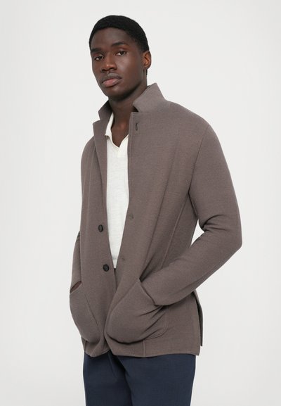 Brun texturerad cardigan med krage, knappstängning och sidofickor, lagd över en ljus skjorta, bärs av en modell.