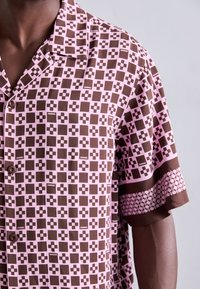 Koszula z krótkim rękawem w brązowo-różowym kolorze z motywem geometrycznym, zapinana na guziki, z kontrastującym wzorzystym wykończeniem wzdłuż rękawów.