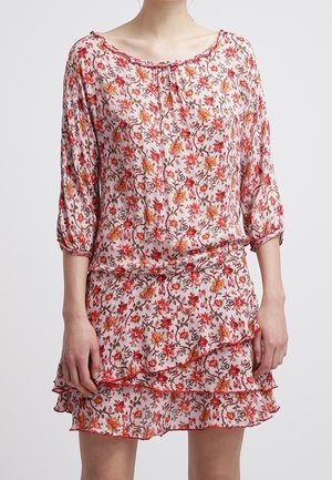 Femme portant une robe rose à fleurs avec des manches trois-quarts et une jupe à volants superposés sur un fond uni.