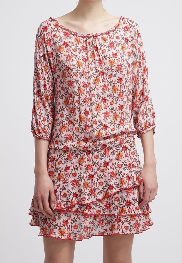 Femme portant une robe rose à fleurs avec des manches trois-quarts et une jupe à volants superposés sur un fond uni.