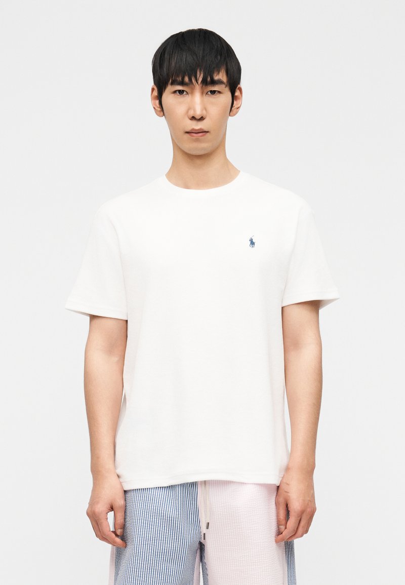 Άνδρας φορώντας απλό λευκό κοντομάνικο T-shirt με μικρό μπλε λογότυπο στο στήθος και ριγέ σορτς, που στέκεται μπροστά σε απλό λευκό φόντο.