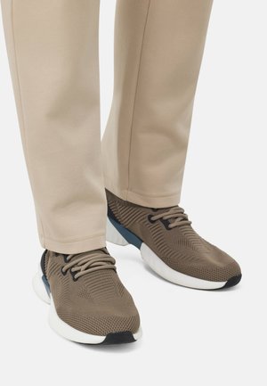Zapatillas deportivas marrones con parte superior de malla texturizada, acentos negros y suela blanca; combinadas con pantalones beige ajustados.