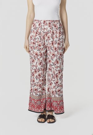 Mujer con pantalones de pierna ancha con estampados florales rojos y un borde decorativo rojo en el dobladillo, combinados con sandalias negras sin talón.