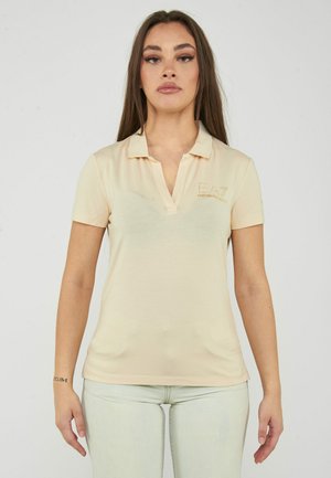 Donna con polo giallo pallido a maniche corte con un piccolo logo sul petto e pantaloni chiari, in piedi davanti a uno sfondo semplice.