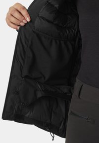 Schwarze Pufferjacke mit einem Innenfutter aus strukturiertem Mesh, Druckknopfverschlüssen, weichem Stoff und einer seitlichen Reißverschlusstasche auf der äußeren Schicht.