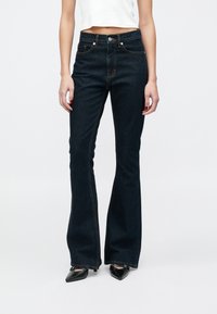 Vaqueros acampanados de color azul oscuro con cintura alta, costuras en contraste, dos bolsillos delanteros y bragueta con cremallera, combinados con zapatos negros de punta fina.