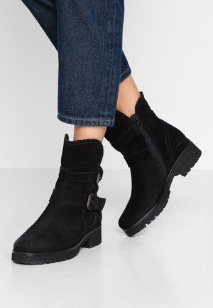 Bottines en daim noir avec fermeture éclair sur le côté et sangles à boucle, portées avec un jean cropped bleu foncé sur un sol clair.