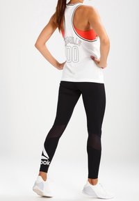 Biały siatkowy top na ramiączkach z czerwonymi akcentami, czarne legginsy z siateczkowymi panelami i białym logo Reebok, w zestawieniu z białymi butami sportowymi.