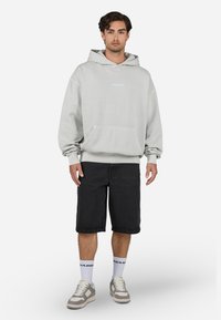 Grå hættetrøje med frontlomme, "PEGAJOR" tekst i lyseblå. Båret med sorte denim shorts, hvide sokker og beige sneakers. Enkel design.