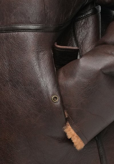 Brązowa skórzana kurtka z miękką podszewką z owczej wełny, widocznymi szwami i detalem z zatrzaskiem. Tekstura jest lekko szorstka.