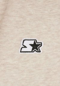 Beige teksturert stoff har en svart og hvit brodert logo med bokstav- og stjernedesign, plassert sentralt.