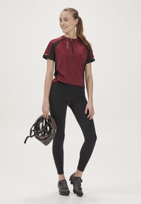 Jersey rowerki z krótkim rękawem w kolorze burgundowym i czarnym z zamkiem, połączony z czarnymi legginsami. Kask rowerowy trzymany w jednej ręce, na nogach czarne buty rowerowe.
