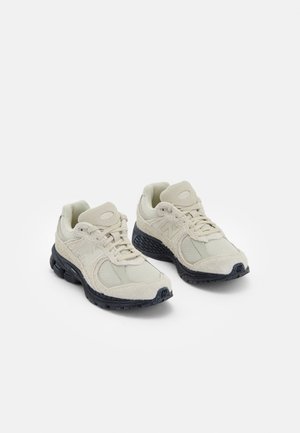 Beżowe sportowe sneakersy z czarnymi podeszwami, siateczkową i zamszową cholewką, sznurowane, pokazane obok siebie na białym tle.