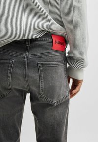 Jeans in denim grigio con un'etichetta rossa "HUGO". Presentano due tasche posteriori e una texture sbiadita, abbinati a un maglione a righe verticali.