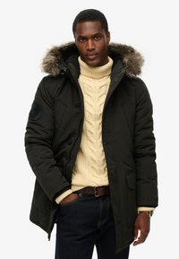 Zwarte gewatteerde parka met een met bont gevoerde capuchon, rits- en knoopsluiting, twee voorkruiszakken, gedragen over een crème kabelbreisel truie en donkere jeans.