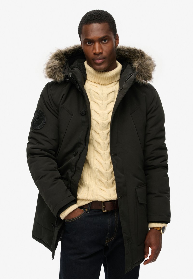 Zwarte gewatteerde parka met een met bont gevoerde capuchon, rits- en knoopsluiting, twee voorkruiszakken, gedragen over een crème kabelbreisel truie en donkere jeans.