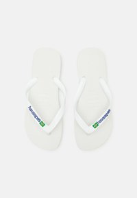 Vita flip-flops i gummi; texturerad fotsula; breda, platta remmar med "havaianas"-logotyp och en accent av den grön-gula brasilianska flaggan.