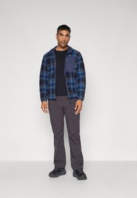 Fleecejakke med blå og svart rutet mønster, glidelås foran og brystlomme; kombinert med mørke bukser og svarte joggesko.