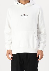 Bílá mikina s kapucí a přední kapsou, s logem "HILFIGER NEW YORK" v námořnické barvě. Hladké manžety a lem, měkká textilní struktura.