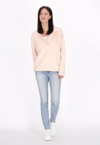 Pullover in tessuto color pesca chiaro con collo a zip a un quarto, progettato con una vestibilità comoda. Abbinato a jeans blu chiaro e scarpe da ginnastica bianche.