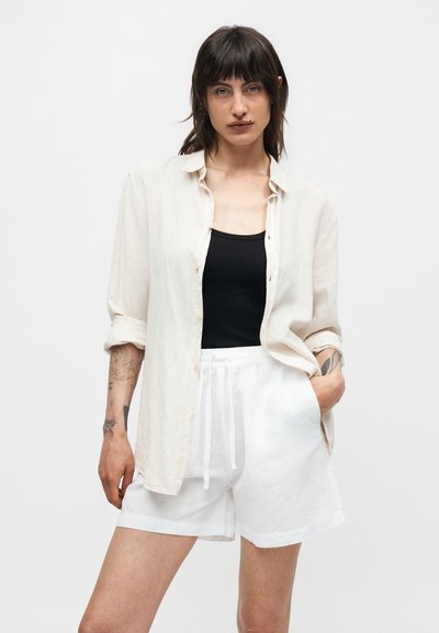 Γυναίκα με σκούρα μαλλιά, που φοράει ένα ανοιχτό μπεζ πουκάμισο πάνω από μαύρο τοπ και λευκά σορτς με κορδόνι, όρθια με το ένα χέρι στην τσέπη.