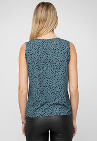 Vrouw met lang blond haar in een mouwloze teal top met zwarte stippen en zwarte leren broek, gezien van achter.