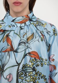 Donna che indossa una blusa blu a collo alto con un motivo floreale che presenta foglie e bacche arancioni, rosa, gialle e verdi.