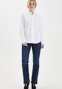 Camicia bianca a maniche lunghe con colletto a punta, abbinata a jeans a gamba dritta blu scuro, con cuciture a contrasto e scarpe con tacco.
