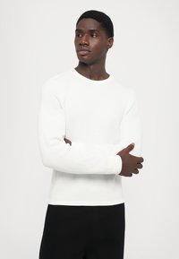 Cămașă albă din tricot cu mâneci lungi, cu guler rotund și textură cu grile, purtată peste pantaloni negri, ținută de un model cu brațele încrucișate.