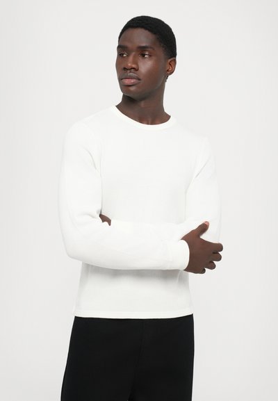 Balts adīts garās piedurknes krekls ar apaļu apkakli un ribotu tekstūru, uzvilkts virs melnajām biksēm, ko tur modelis ar apskāvušām rokām.