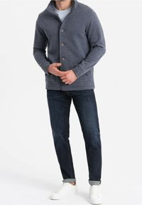 Šedý cardigan na knoflíky s vysokým límcem, vyrobený z měkkého materiálu. Nesený přes světlou košili a kombinovaný s tmavými džíny a bílými teniskami.