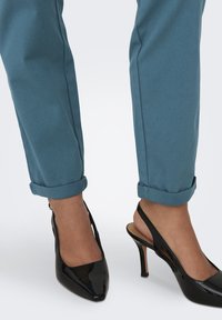 Melnas patentā ādas augstpapēžu kurpes ar smailu purngalu un izgriezumiem, savienotas ar sarullētām teāla krāsas auduma biksēm.