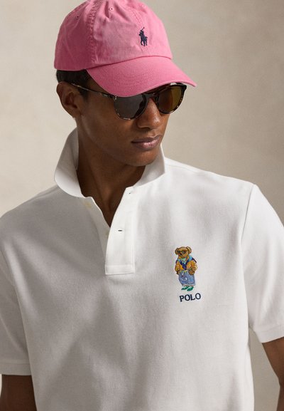 Mann iført briller med skilpaddemønster, rosa caps og hvit polotrøye med brodert bjørnelogo og teksten "POLO".