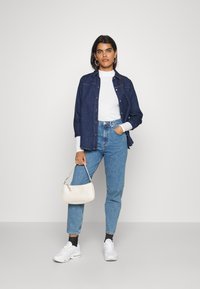 Ung kvinne står med hvit håndveske, iført hvit genser med høyt hals, mørk denimskjorte, blå jeans, hvite sneakers og store hoopear. 