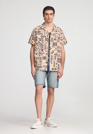 Camicetta a maniche corte beige con motivi astratti marroni e neri, abbinata a pantaloni shorts in denim. Le calzature presentano una combinazione di colori crema e bianco.