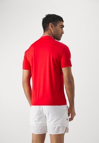 Maglietta sportiva rossa con collo a polo, maniche corte e texture liscia, abbinata a pantaloni corti bianchi con un piccolo logo.