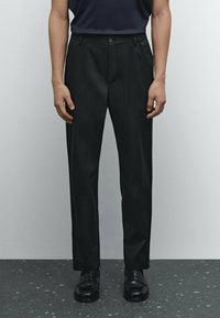 Black tailored trousers featuring a slim fit, flat front design, and a smooth texture. Reflective of minimalist style with a classic button closure.