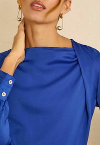 Modrá blúzka s hladkou textúrou, lodičkovým výstrihom a zhladeným detailom na pleciach. Dlhé rukávy s gombíkmi na manžetách. Zlaté a strieborné náušnice.