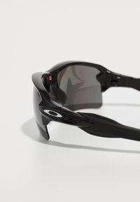 Černé sportovní sluneční brýle s tmavými čočkami, aerodynamický design a hladký plastový rám. Na straně se nachází detail loga.