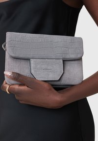 Grå ruskinds clutch med et tekstureret krokodillemønster, der har en klaplukning og en rektangulær forlommer. Minimalistisk design med diskret branding.