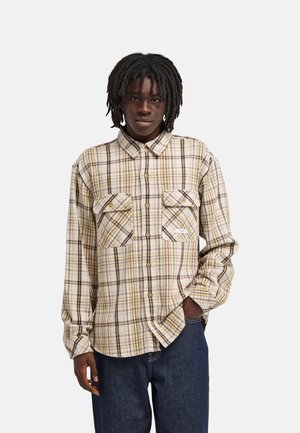 Young man with dreadlocks wearing a beige plaid button-up shirt with pockets and dark blue jeans standing against a plain background.