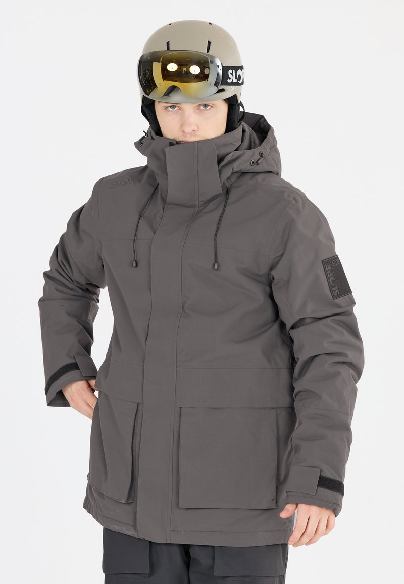 Grå vandtæt jakke med høj krave, justerbare snore, frontlommer og sorte velcro-detaljer, båret med en let hjelm og beskyttelsesbriller.
