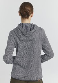 Huppari mustavalkoisessa geometrisessa kuosissa, jossa on pitkät hihat, teksturoitu kangas ja kiinteä musta reunus hupun ympärillä.