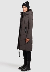 Vrouw in een lange bruine gewatteerde jas met capuchon, zwarte beanie, zwarte leggings en zwarte laarzen, staande in profiel tegen een effen achtergrond.