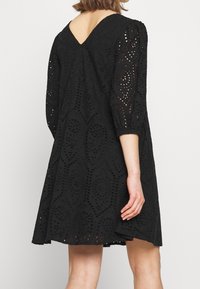 Musta reikäkoristeinen mekko, jossa on levenevä muotoilu, kolmanneksi viimeiset hihat ja V-muotoinen selkäosa, sekä monimutkaisia rei'itettyjä kuvioita koko mekossa.