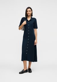 Μεσαίο φόρεμα σε ναυτικό μπλε, με κοντά μανίκια, λεία υφή και χρυσά κουμπιά. Το μοντέλο κρατά μια τσάντα με σχέδια και φορά μαύρα μοκασίνια.