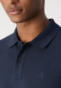 Marine blå polo skjorte med teksturert stoff, med krage, to svarte knapper og en liten brodert logo på brystet.
