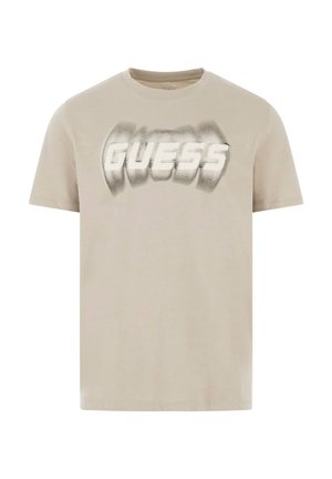 Svetlo bež majica iz bombaža s zamegljenim logotipom "GUESS" v beli in sivi barvi. Kratki rokavi in okroglo izrezan izrez.